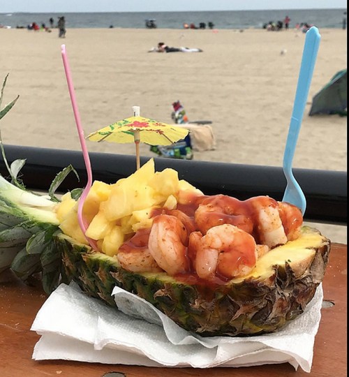 A pineapple boat filled with shrimp cocktail, garnished with an umbrella, on a beach with people in the background.