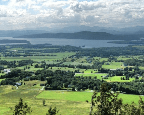 A panoramic view of lush green fields and a lake surrounded by mountains under a partly cloudy sky.