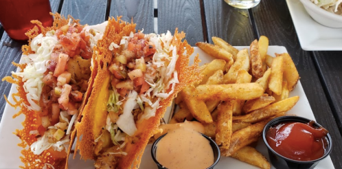 Two crispy tacos filled with toppings, served with seasoned fries and dipping sauces on a white plate.