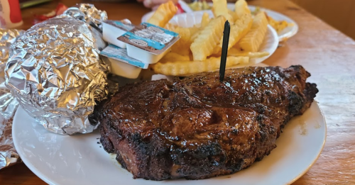A plate with a large grilled steak, a foil-wrapped potato, and crinkle-cut fries with dipping sauces.