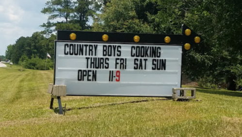 Sign for "Country Boys Cooking" with hours: Thurs-Sun, 11 AM to 9 PM, located along a grassy roadside.