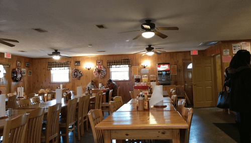 Cozy restaurant interior with wooden walls, tables, and fans; patrons dining and a drink station in the background.