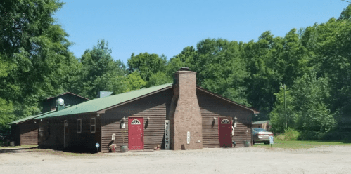 A rustic wooden building with red doors, surrounded by trees and a gravel driveway.