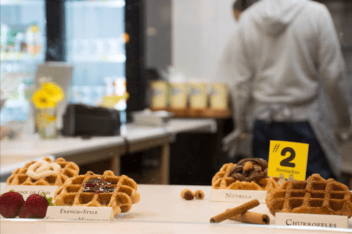 A display of various waffles, including French-style, Nutella, and churro flavors, with a blurred figure in the background.