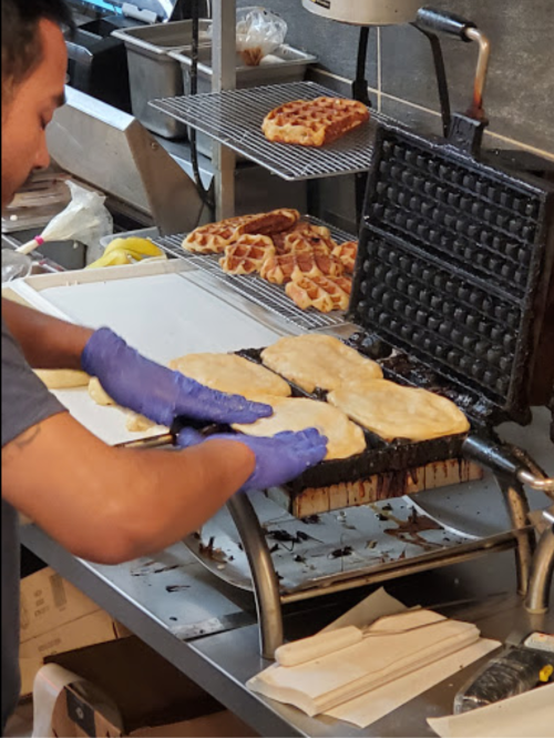 A person in gloves prepares dough on a grill in a busy kitchen, with baked goods in the background.