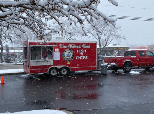 A red food truck named "The Killed Cod" serves fish and chips in a snowy parking lot. Snowflakes are falling.