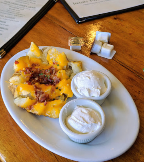 A plate of cheesy baked potatoes topped with bacon bits, served with two cups of sour cream on the side.