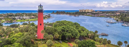 Aerial view of a red lighthouse surrounded by lush greenery and a blue river, with buildings in the distance.