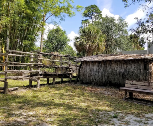 A rustic thatched hut beside a wooden fence and a bench, surrounded by trees and blue skies.