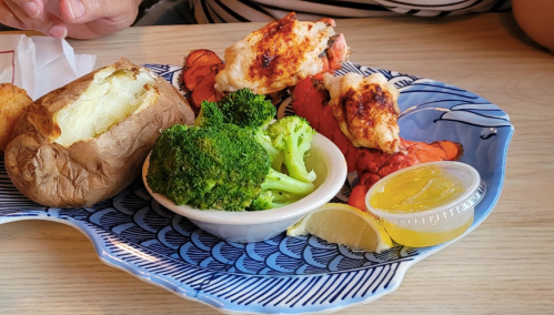 A plate with two lobster tails, a baked potato, steamed broccoli, and a lemon wedge, served with a dipping sauce.