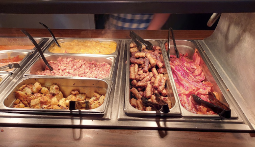 A buffet display featuring various cooked meats, including sausages, bacon, and seasoned potatoes in metal trays.