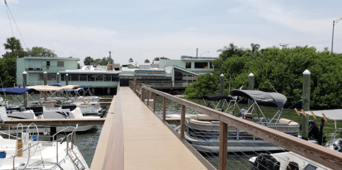 A wooden dock leads to a marina with boats and green foliage in the background under a clear sky.