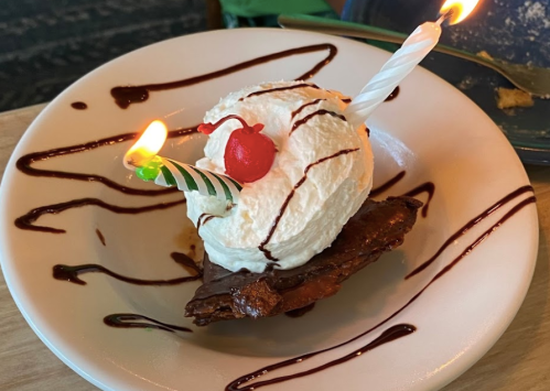 A slice of brownie topped with whipped cream, a cherry, and two lit birthday candles on a plate drizzled with chocolate sauce.