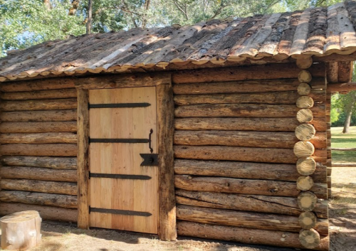 A rustic log cabin with a wooden door and a slanted roof, surrounded by trees in a natural setting.