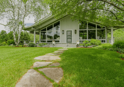 A modern house with large windows surrounded by lush greenery and a stone pathway leading to the entrance.