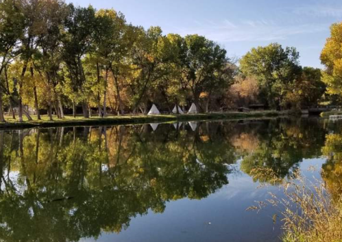 A serene landscape featuring a calm pond reflecting trees and teepees under a clear blue sky.