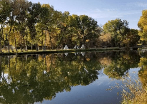 A serene landscape featuring a calm pond reflecting trees and teepees under a clear blue sky.