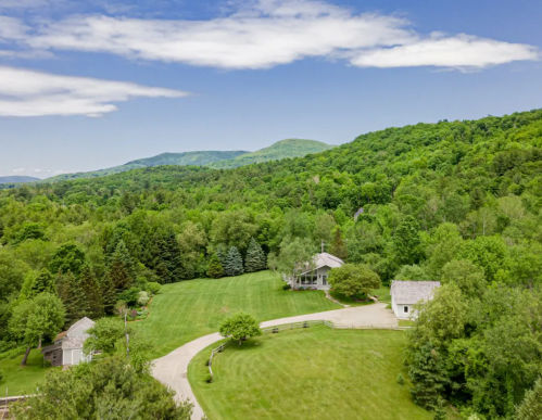 Aerial view of a lush green landscape with a house and barn, surrounded by trees and rolling hills under a blue sky.