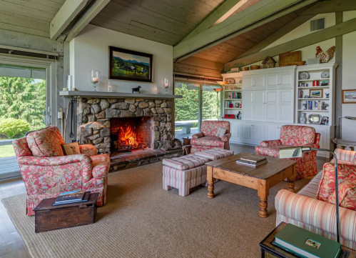 Cozy living room with a stone fireplace, floral armchairs, a wooden coffee table, and large windows overlooking greenery.
