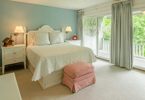 Bright bedroom with a white bed, light blue walls, and large windows overlooking greenery. Cozy decor with a pink ottoman.