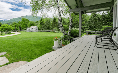 A peaceful porch view overlooking a green lawn, trees, and a distant shed against a backdrop of mountains.
