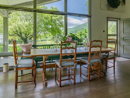 A spacious dining area with a wooden table, chairs, and large windows showcasing a scenic view of greenery and mountains.