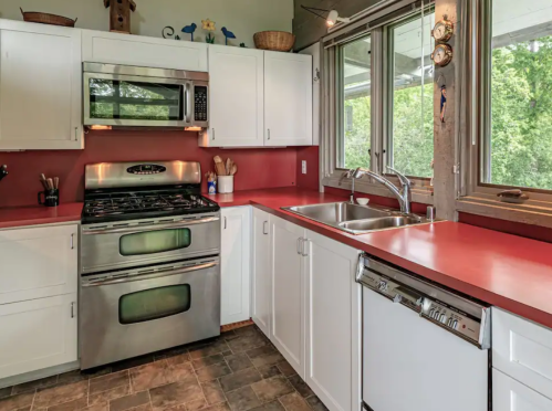 Modern kitchen with stainless steel appliances, red countertops, white cabinets, and large windows overlooking greenery.