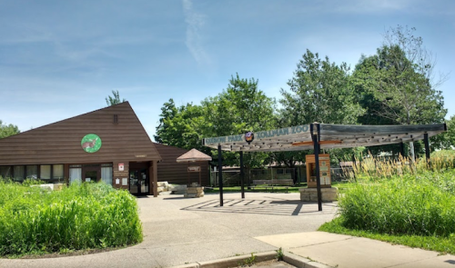 Entrance to a zoo with a wooden building and a sign, surrounded by greenery and trees on a sunny day.