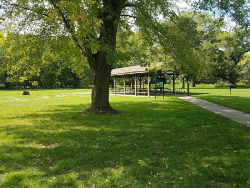 A grassy park area featuring a shaded pavilion and trees under a clear blue sky.