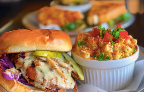 A crispy chicken sandwich with pickles and slaw, served alongside a bowl of creamy macaroni and cheese topped with tomatoes.