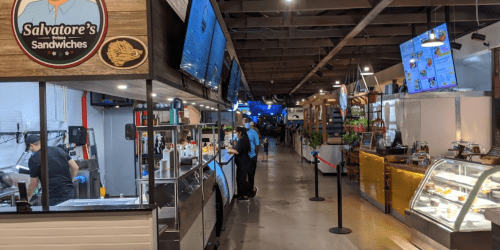 A bustling food hall with various vendors, including Salvatore's Sandwiches, and customers ordering food.