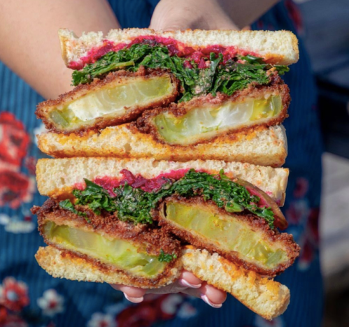 A person holds a stacked sandwich filled with crispy pickles, greens, and vibrant pink sauce between slices of bread.