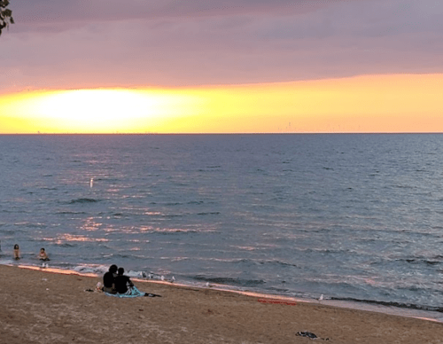 A couple sits on the beach at sunset, with waves gently lapping at the shore and a colorful sky in the background.