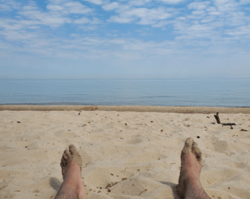 A pair of bare feet resting on sandy beach, with calm water and a cloudy sky in the background.