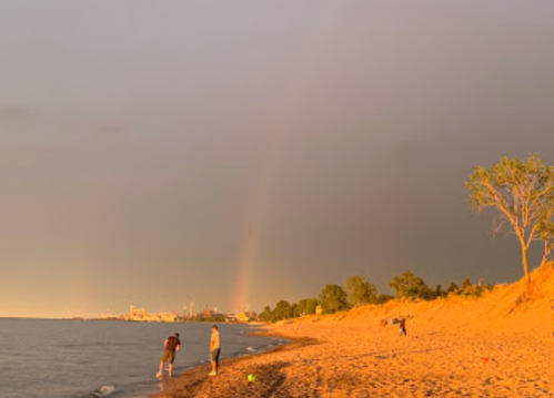 A beach scene at sunset with a rainbow in the background and two people walking along the shore.