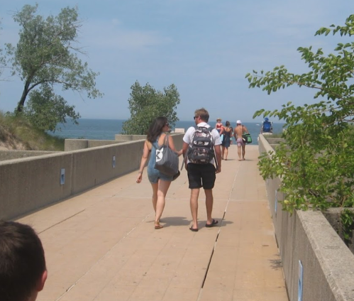 A couple walks hand in hand along a pathway by the water, with trees and other people in the background.