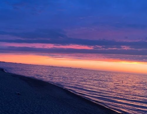 A serene beach at sunset, with vibrant purple and orange hues reflecting on calm water under a cloudy sky.