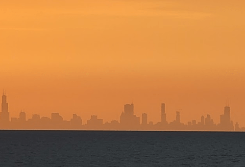 Silhouette of a city skyline against an orange sunset, viewed over calm waters.
