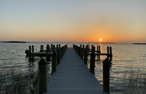A serene sunset over a calm waterway, viewed from a wooden pier lined with posts.