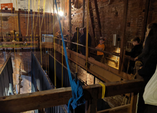 A group of people stands on a wooden platform overlooking a large, deep space with ropes and industrial features.