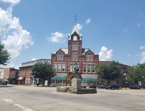 Historic brick building with a clock tower, surrounded by trees and a statue in the foreground, under a blue sky.