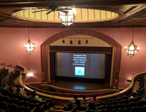 Interior of a theater with a screen displaying "Support the businesses that support your local community" and audience seated.