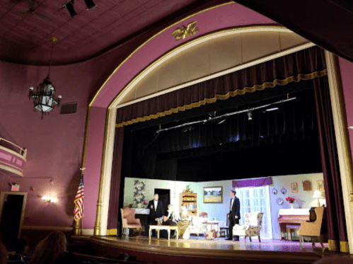 A theater stage with a pink backdrop, featuring a living room set and two actors performing a scene.