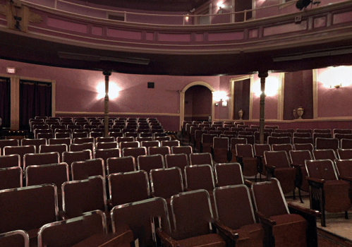Interior of a theater with empty rows of red seats and a softly lit stage area in the background.