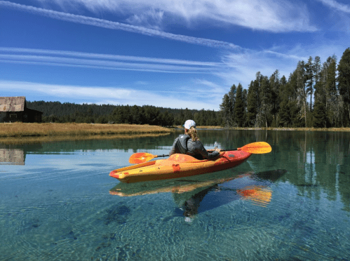 A person kayaking on a clear lake surrounded by trees and a blue sky with wispy clouds.