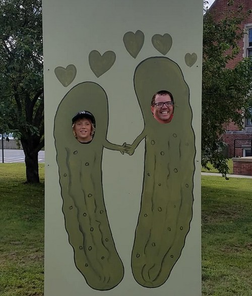 Two people with their faces in a pickle-themed cutout, holding hands with hearts above them.