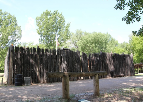 A wooden fort wall surrounded by trees, with a bench in the foreground and a clear blue sky above.