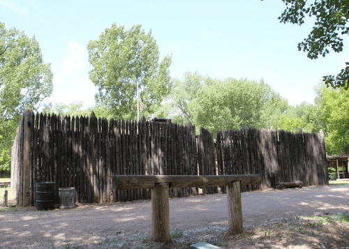 A wooden fort wall surrounded by trees, with a bench in the foreground and a clear blue sky above.