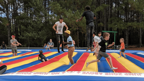 A group of children and teens jumping and playing on a colorful inflatable bounce pad in a wooded area.