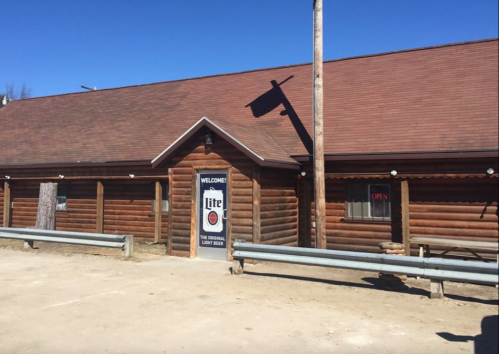 Log cabin-style building with a "Welcome" sign and an "Open" sign, surrounded by gravel and a clear blue sky.